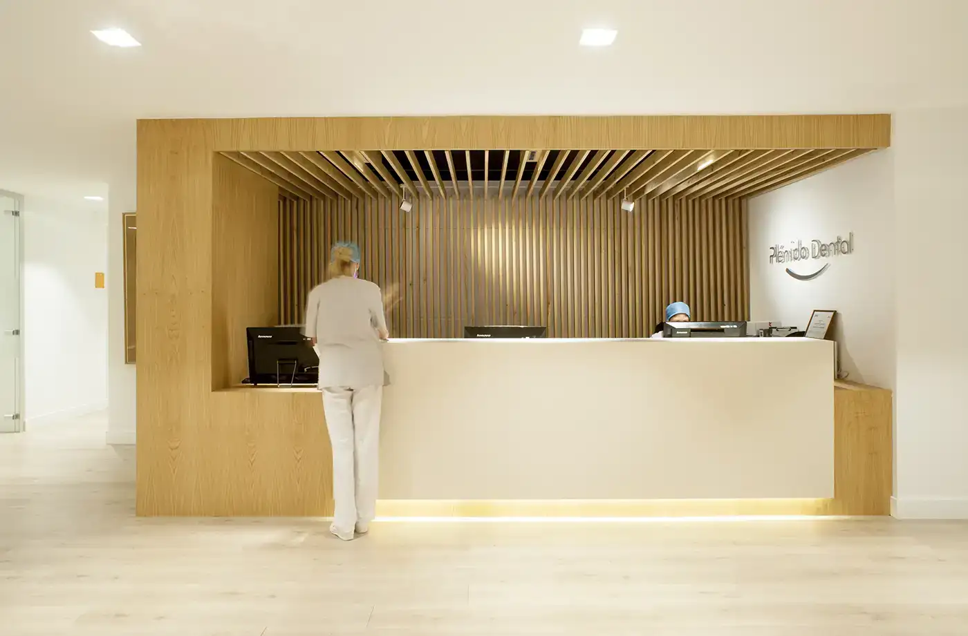 Interior clínica dental. Vista mostrador. Iluminación cálida, tonos crema y madera.