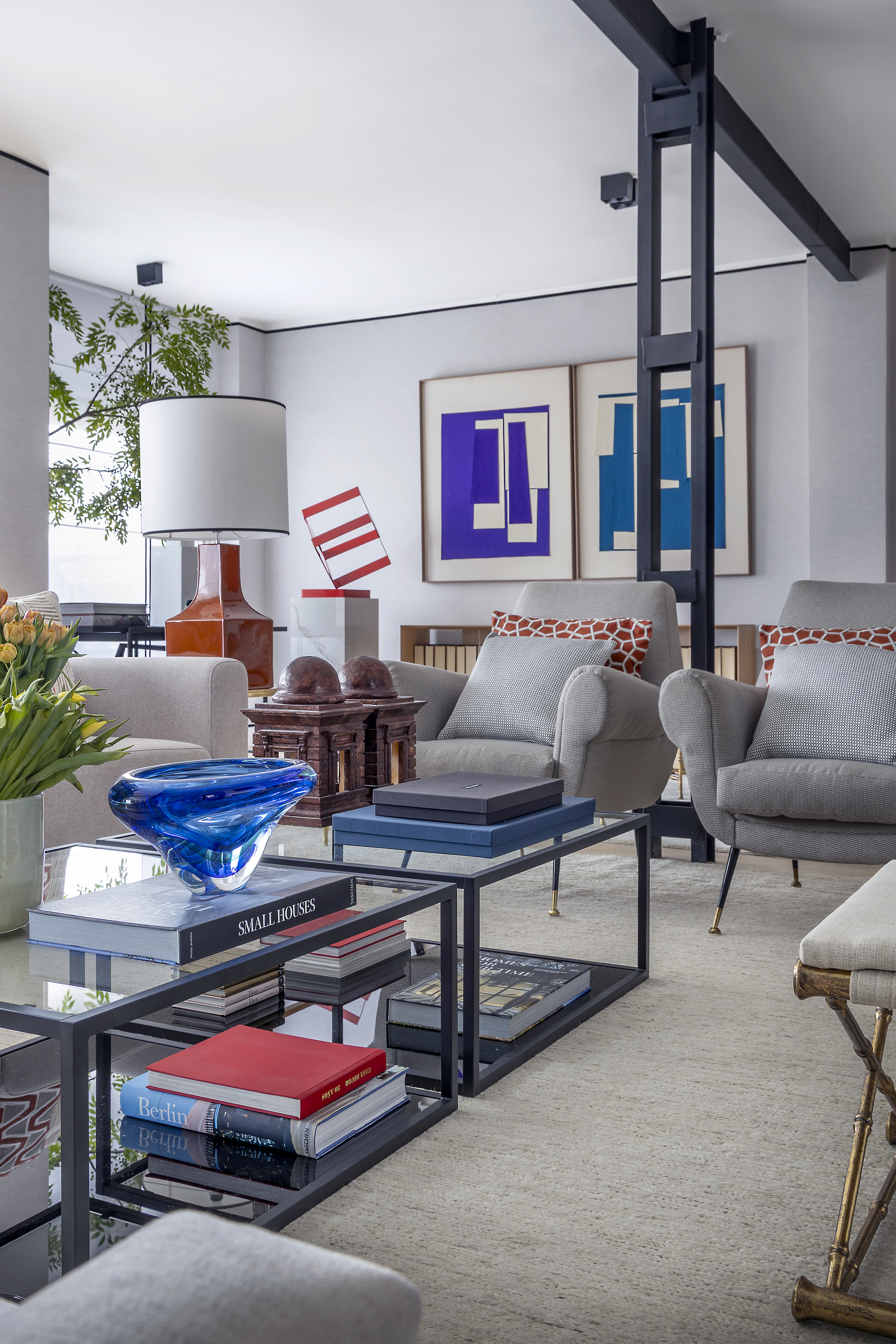 Salón moderno con mesas de centro, libros y elementos decorativos, butacas tapizadas y obras de arte en la pared.