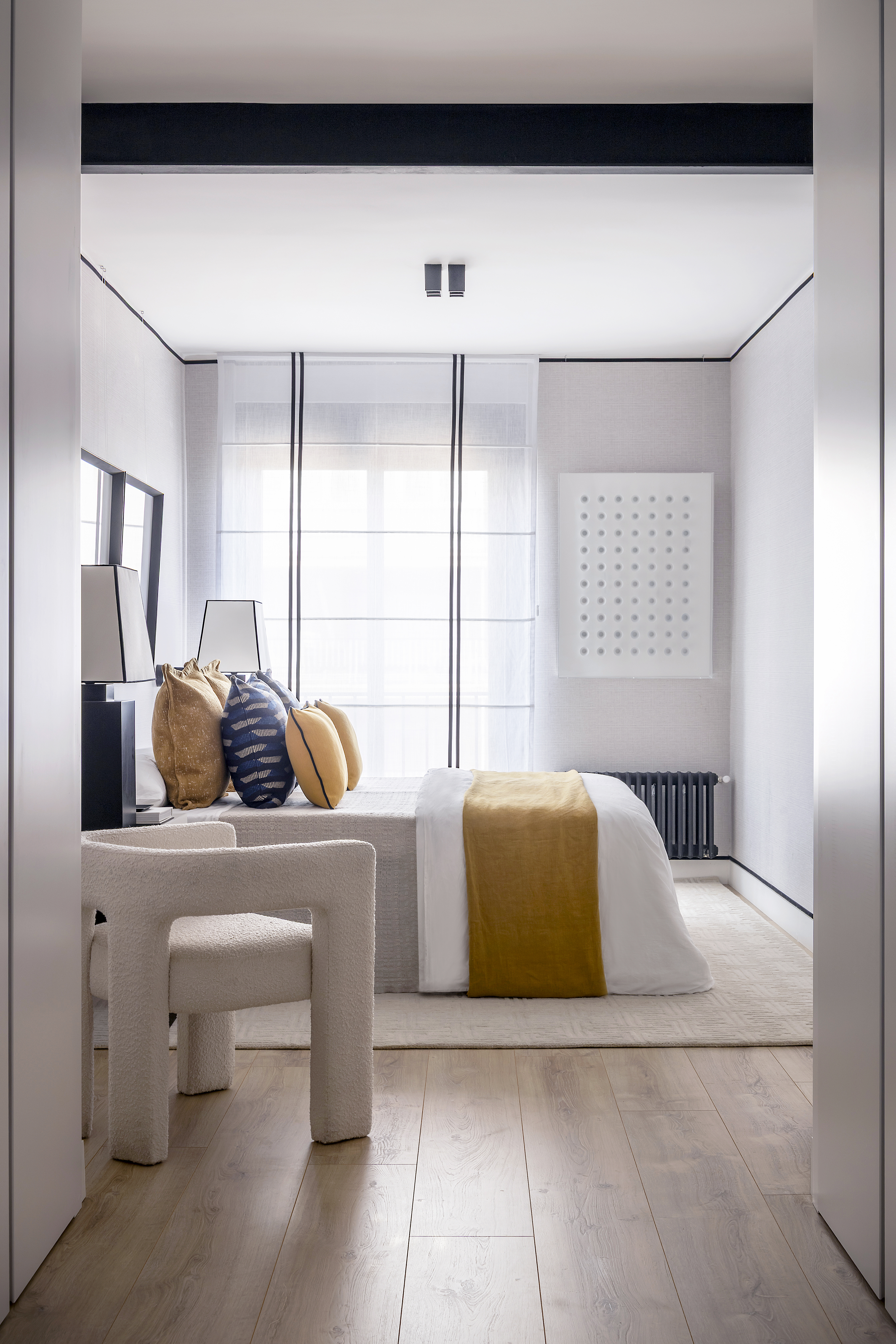 Vista del dormitorio con cama vestida, sillón auxiliar y entrada de luz natural a través de la ventana.