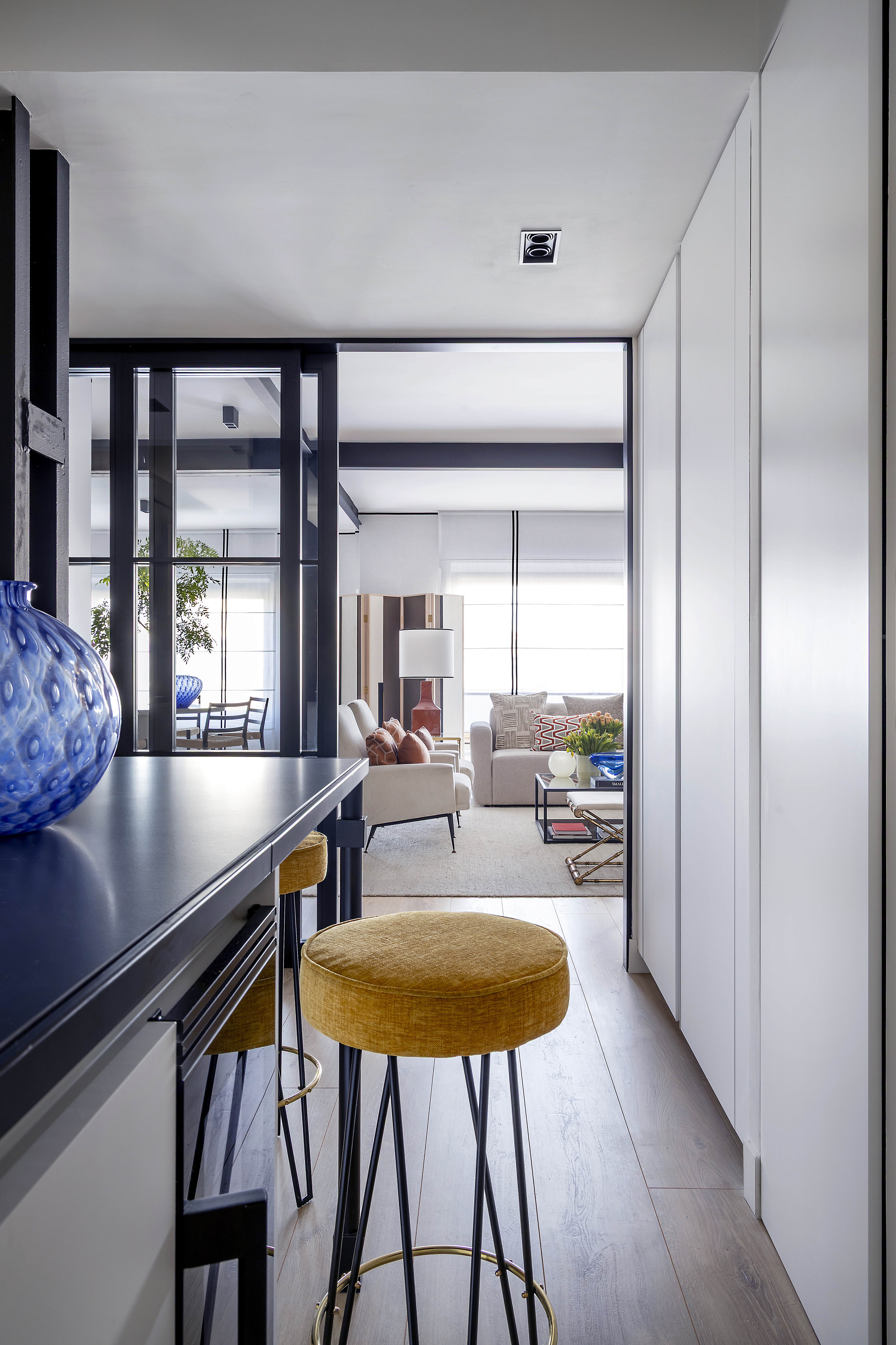 Vista desde la cocina hacia el salón a través de puerta corredera acristalada, con barra y taburetes en primer plano.