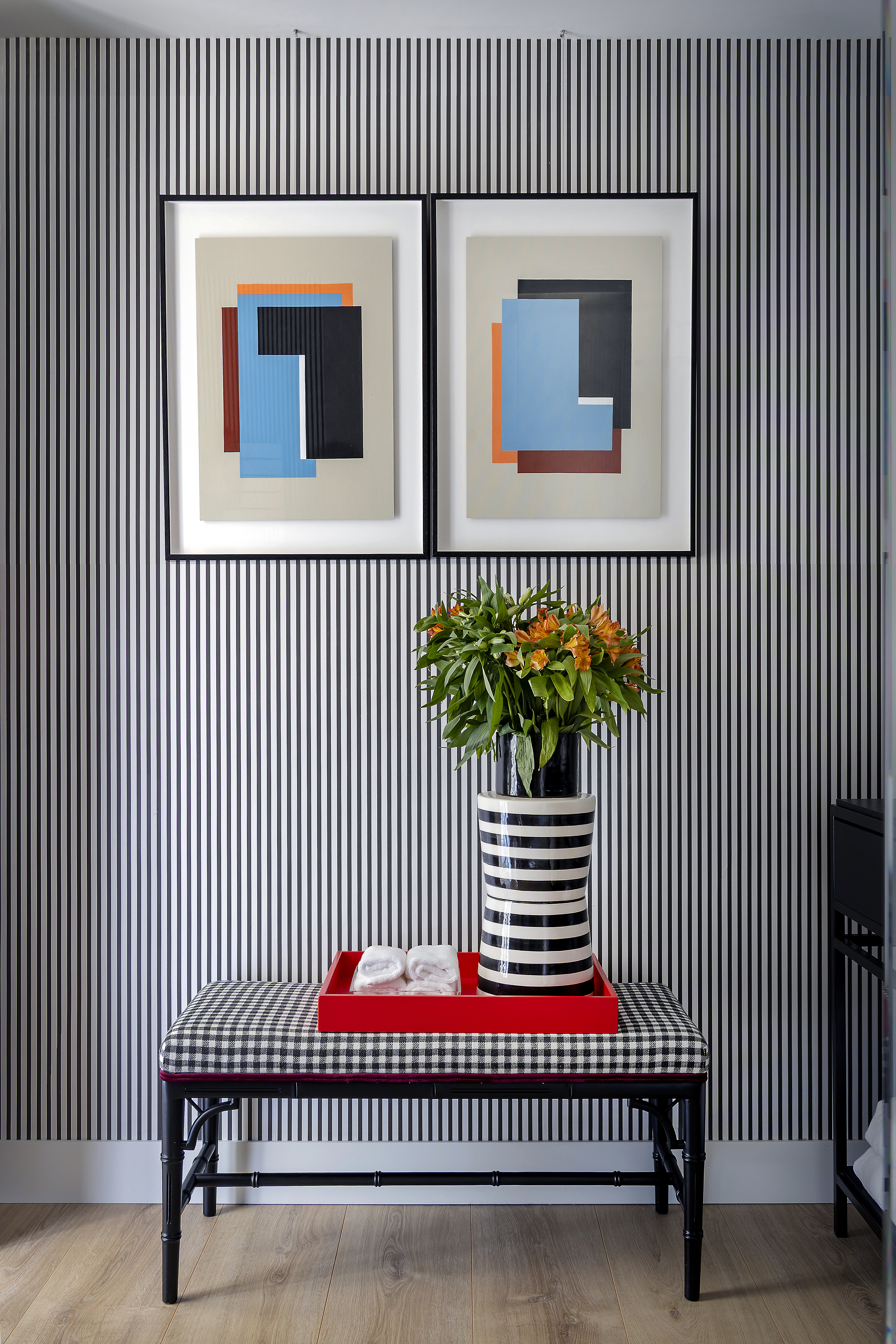 Detalle decorativo del baño con banco auxiliar, jarrón con flores y obras de arte en la pared.