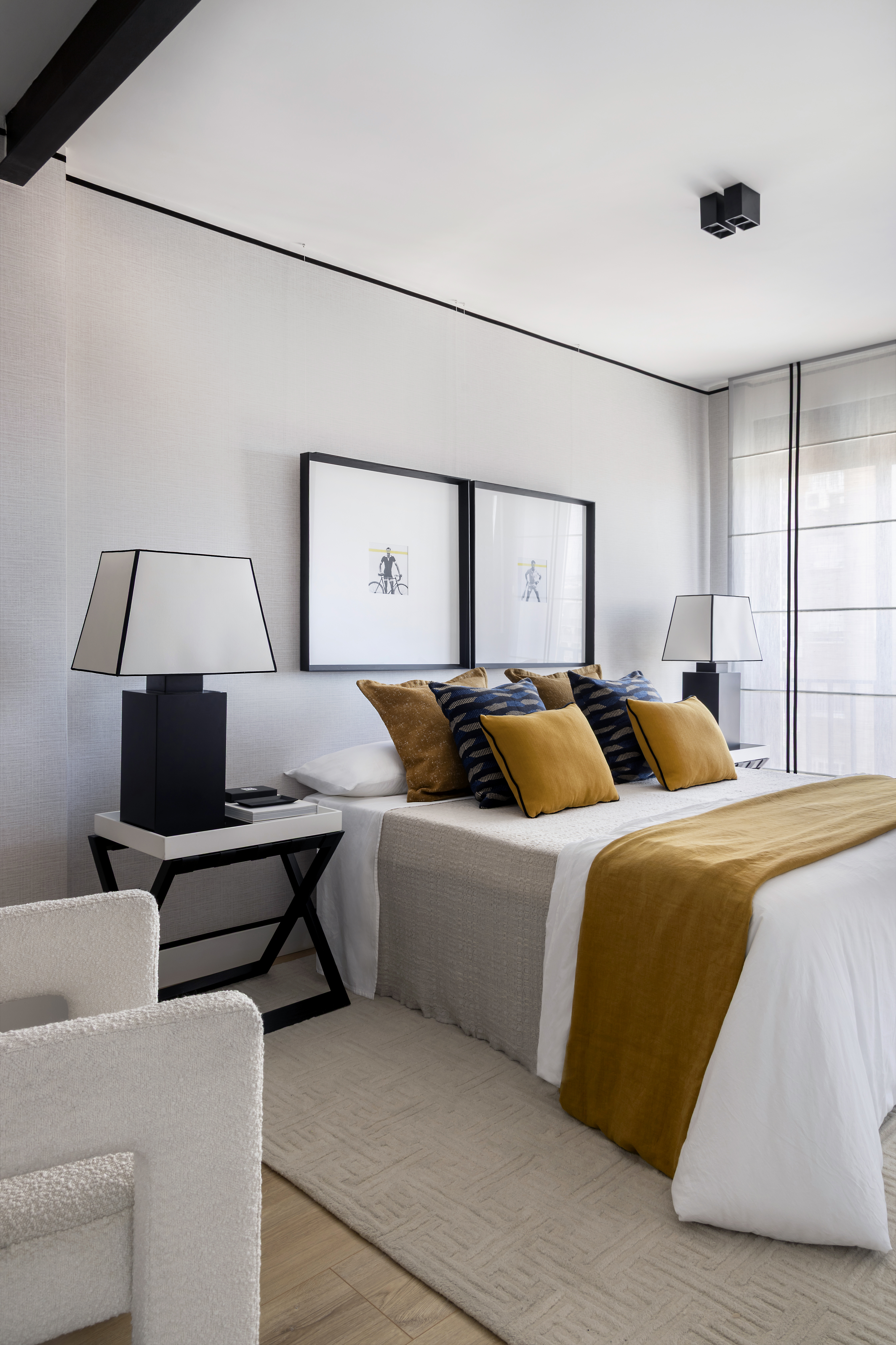 Vista del dormitorio con cama vestida, mesillas con lámparas, textiles decorativos y sillón auxiliar.